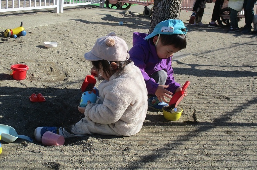 1歳児が園庭で砂遊び。スコップで砂をすくい、カップへ入れようとしている。