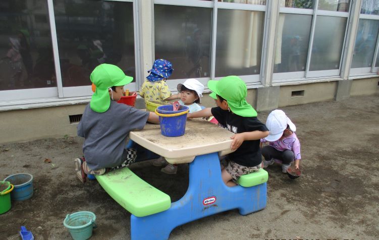 子どもたちが園庭にあるイステーブルに座り、バケツとスコップを使ってままごと遊びをしている。